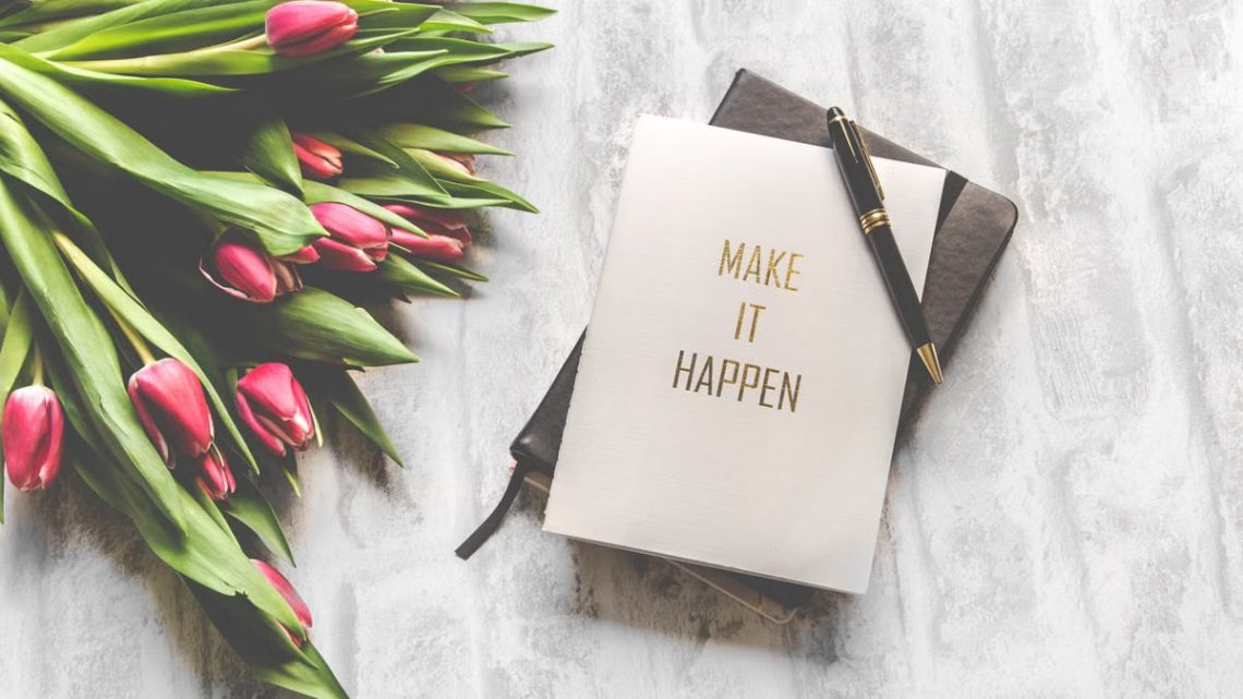A notebook with 'Make It Happen' inscription beside a bouquet of pink tulips, capturing inspiration.